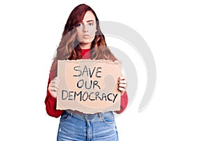 Young beautiful woman holding save our democracy protest banner thinking attitude and sober expression looking self confident