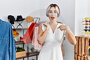 Young beautiful woman holding shopping bags at retail shop pointing finger to one self smiling happy and proud