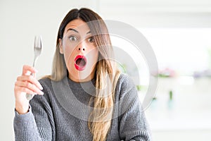Young beautiful woman holding silver fork at home scared in shock with a surprise face, afraid and excited with fear expression