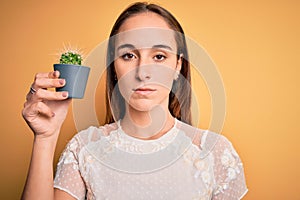 Young beautiful woman holding small cactus plant pot over  yellow background with a confident expression on smart face