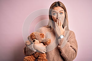 Young beautiful woman holding teddy bear standing over isolated pink background cover mouth with hand shocked with shame for