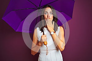 Young beautiful woman holding umbrella standing over purple isolated background cover mouth with hand shocked with shame for