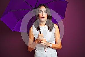 Young beautiful woman holding umbrella standing over purple isolated background scared in shock with a surprise face, afraid and