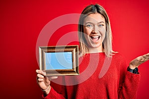 Young beautiful woman holding vintage frame standing over isolated red background very happy and excited, winner expression