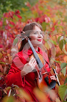 Young beautiful woman holding violin among fall colored red and green leaves in autumn