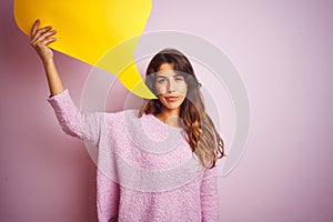 Young beautiful woman holding yellow speech bubble standing over pink isolated background with a confident expression on smart