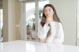 Young beautiful woman at home on white table looking confident at the camera with smile with crossed arms and hand raised on chin