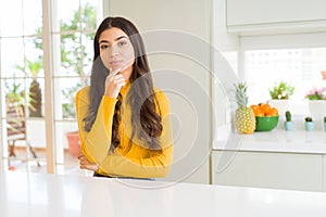 Young beautiful woman at home on white table looking confident at the camera with smile with crossed arms and hand raised on chin