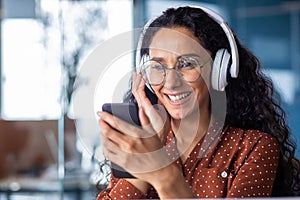 Young beautiful woman inside office with phone in hands and headphones listening to music and watching online video