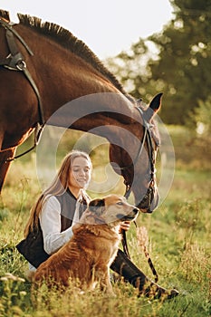 A young beautiful woman jockey with her dog sits in a meadow near her horse at sunset.