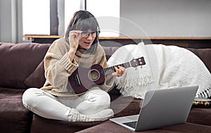 Young beautiful woman learning to play ukulele at home with online lessons