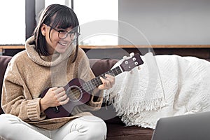 Young beautiful woman learning to play ukulele at home with online lessons