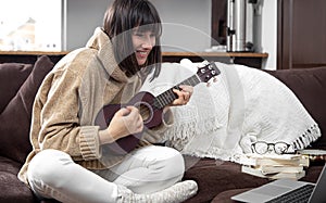 Young beautiful woman learning to play ukulele at home with online lessons