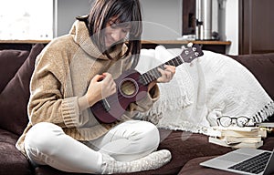 Young beautiful woman learning to play ukulele at home with online lessons