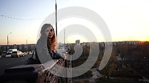 A young beautiful woman in a leather jacket with a black backpack stands thoughtfully on the bridge against the