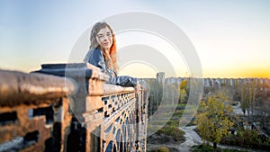 A young beautiful woman in a leather jacket with a black backpack stands thoughtfully on the bridge against the