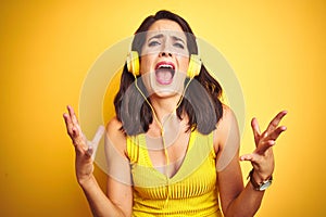 Young beautiful woman listening to music using headphones over yellow isolated background crazy and mad shouting and yelling with