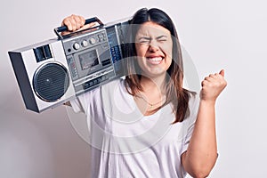 Young beautiful woman listening to music using vintage boombox over white background screaming proud, celebrating victory and