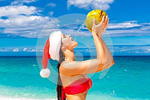 Young beautiful woman with long black hair in red bikini, dressed in red Santa Claus hat is drinking coconut water on tropical be