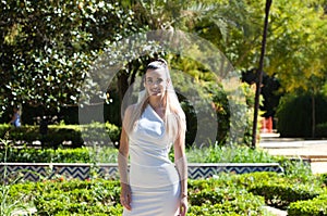 Young and beautiful woman with long blond hair and a short white dress is in a famous park in seville rich in vegetation, spain.