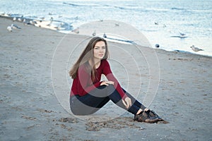 young beautiful woman with long hair, in black jeans and red shirt, sitting on sand on beach among seagulls birds