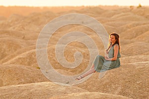 Young beautiful woman with long hair in blue dress sitting elegant and thoughtful on sand at sunrise in sandy desert