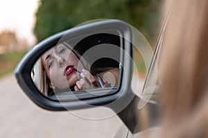 A young, beautiful woman with long hair through the car window looks in the rear view mirror and paints her lips