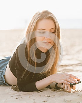 Young beautiful woman with long hair is relaxing on sandy beach with mobile phone in backlight.