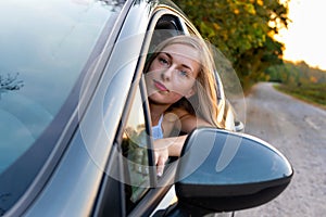 A young, beautiful woman with long hair sits at the wheel of the car and looks out the window