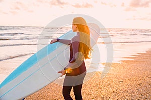 Young beautiful woman with long hair. Surf girl in wetsuit with surfboard on a beach at sunset or sunrise.