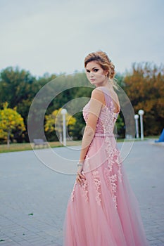 Young beautiful woman in long pink evening dress walking path in park. Fashion style portrait of gorgeous beautiful girl outdoors