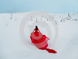 Young beautiful woman in long red dress over winter hill background and snowfall. Fairy tale girl on polar winter landscape. View