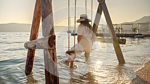 Young beautiful woman in long white dress relaxing on the rope swing at sea beach lit by sunset rays. Tourism