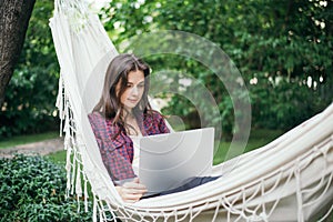 Young beautiful woman lying in a hammock with laptop in a garden. green background. Trees. telework. remote work