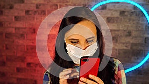 Young beautiful woman in medical mask stands with mobile phone in hands on background brick wall with glowing lamps. Portrait of c