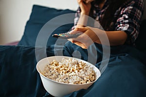 Young beautiful woman in morning bed at home. Hold remote control and watch tv or movie. Bowl with popcorn on bed. Cut