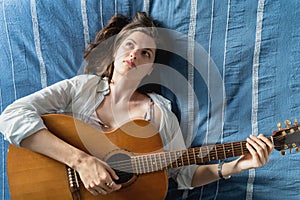 Young beautiful woman, musician, singer studying, practicing to play acoustic five-string guitar at home lying on bed in blue