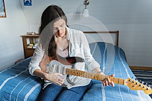 Young beautiful woman, musician, singer studying, practicing to play electric guitar at home sitting on bed in blue shirt,jeans.