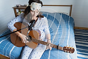 Young beautiful woman, musician, singer studying to play acoustic five-string guitar at home sitting on bed in blue pajamas,