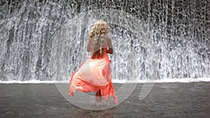 Young beautiful woman in orange dress approaching to big waterfall