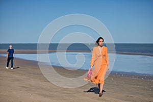 Young and beautiful woman in an orange dress and pink heels in her hand, walking barefoot on the beach in solitude. Concept beauty