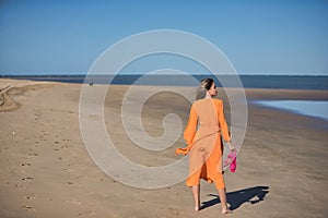 Young and beautiful woman in an orange dress and pink heels in her hand, walking barefoot on the beach in solitude. Concept beauty