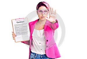 Young beautiful woman with pink hair holding clipboard with contract document doing stop sing with palm of the hand