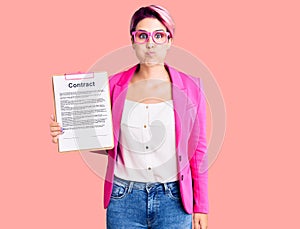 Young beautiful woman with pink hair holding clipboard with contract document puffing cheeks with funny face