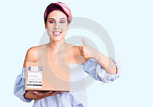 Young beautiful woman with pink hair holding delivery package smiling happy and positive, thumb up doing excellent and approval