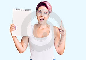 Young beautiful woman with pink hair holding notebook surprised with an idea or question pointing finger with happy face, number