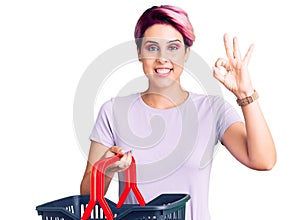 Young beautiful woman with pink hair holding supermarket shopping basket doing ok sign with fingers, smiling friendly gesturing