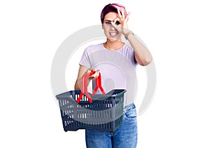 Young beautiful woman with pink hair holding supermarket shopping basket smiling happy doing ok sign with hand on eye looking