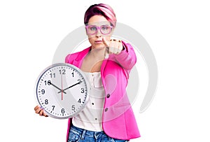 Young beautiful woman with pink hair wearing business jacket and holding clock pointing with finger to the camera and to you,