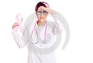 Young beautiful woman with pink hair wearing doctor stethoscope holding cancer ribbon stressed and frustrated with hand on head,